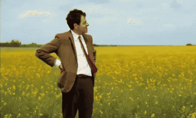 Mr. Bean standing in an empty field, checking his watch and waiting.